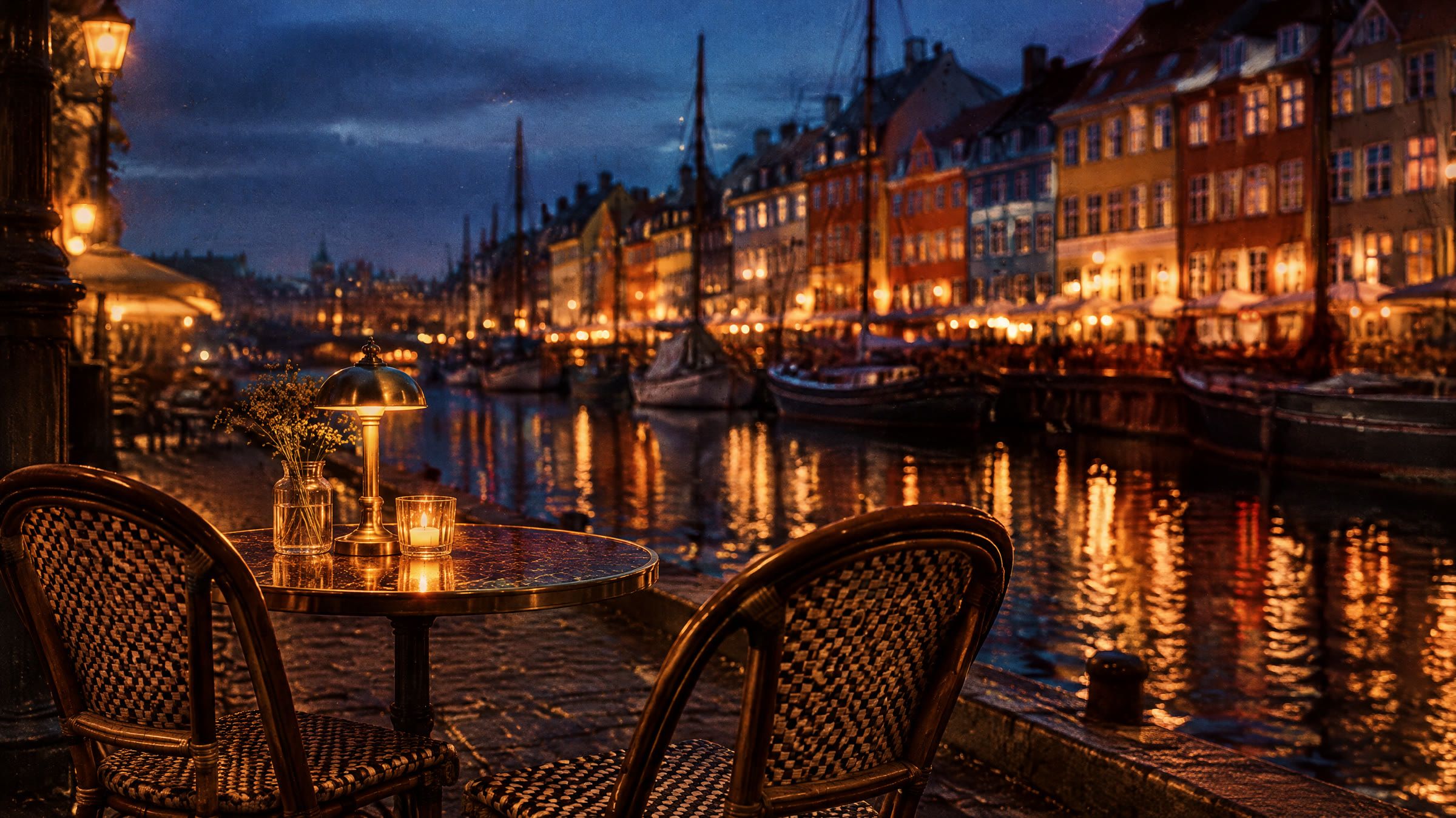 En kanal i København i blå time med farvede gavlhuse spejlet i vandet og to tomme cafestole på kajen.